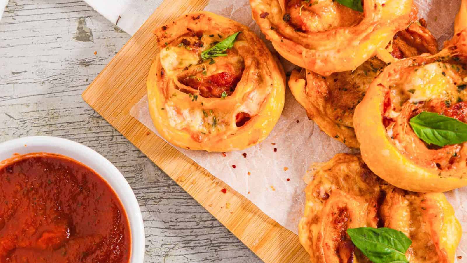 A wooden board holds several pizza rolls topped with basil leaves, set on parchment paper. A small bowl of marinara sauce is on the left. The table surface is light gray with visible grain patterns.
