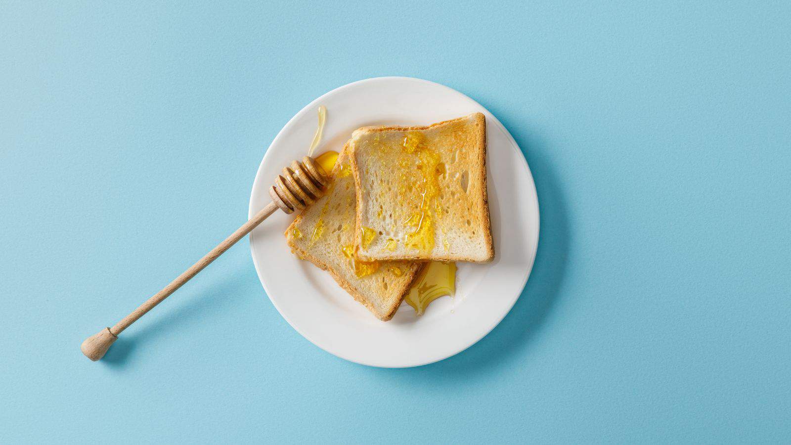 Two slices of toast with honey on a white plate, set against a light blue background. A honey dipper with traces of honey is placed beside the toast on the plate.