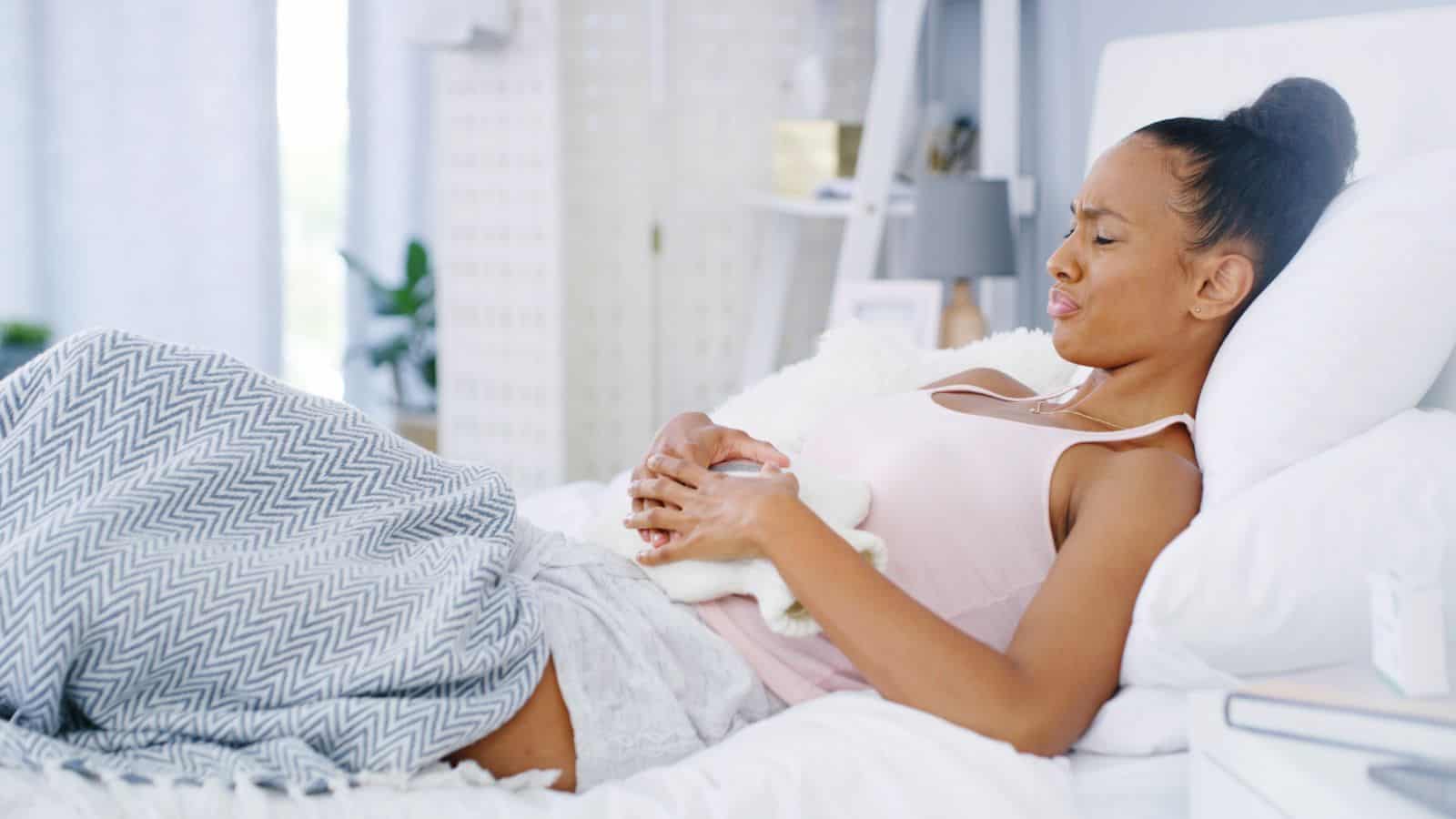 A woman lies on a bed, wearing a pink tank top and gray patterned blanket. She clutches her stomach with a pained expression, holding a hot water bottle. The room is softly lit with a white background and minimal decor.