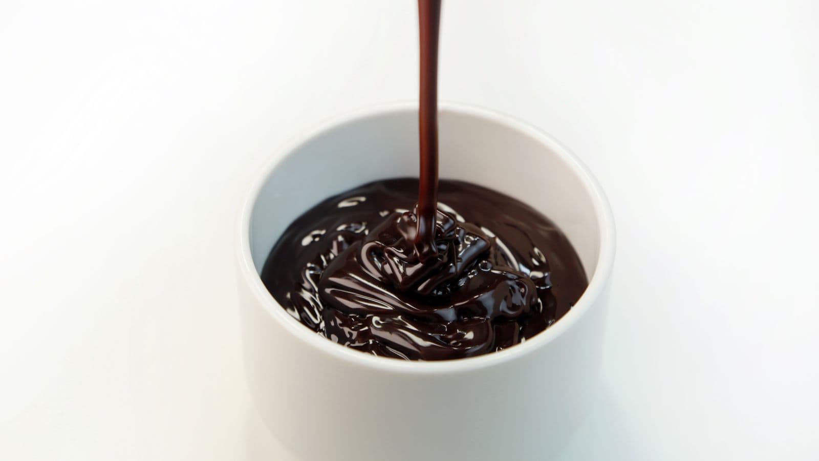 A smooth stream of chocolate sauce is being poured into a white bowl against a plain background.