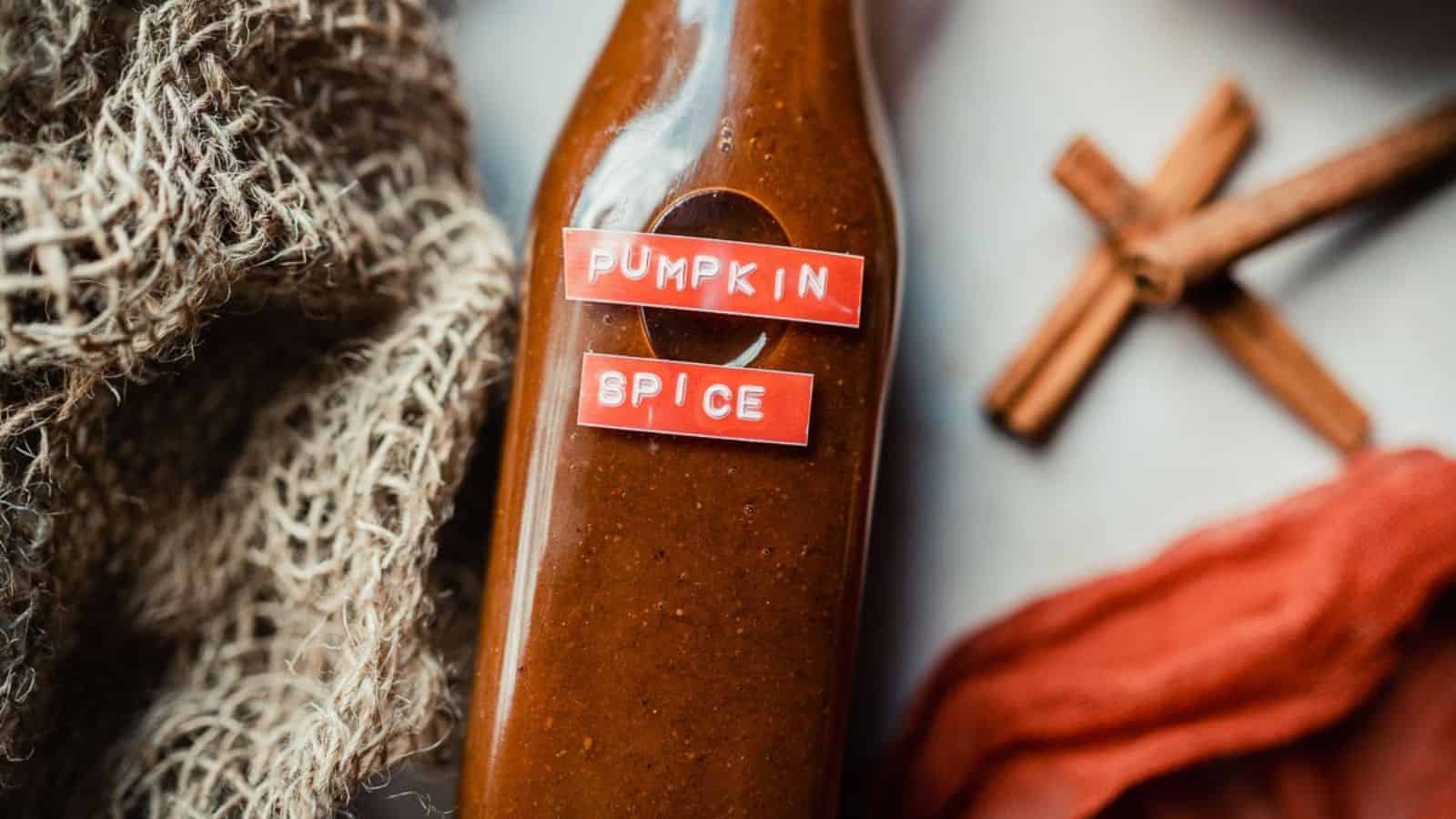 A bottle labeled "Pumpkin Spice" with a reddish-brown liquid inside is placed on a surface. Nearby are textured fabric, a stick of cinnamon crossed with another, and a piece of orange material.