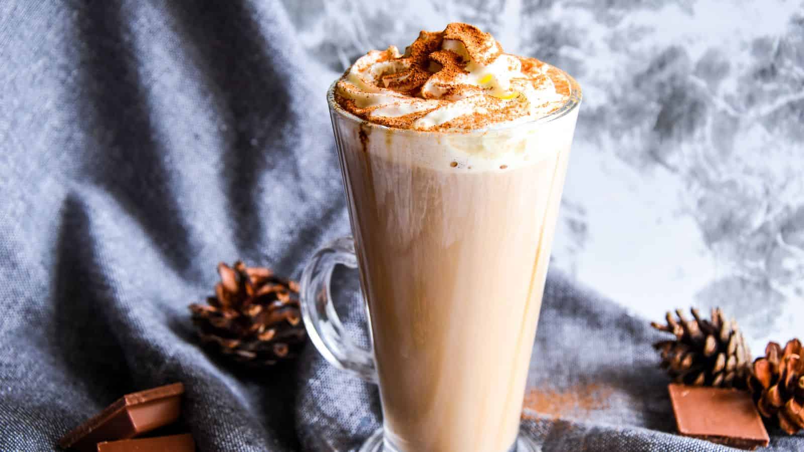 A glass mug filled with frothy coffee topped with whipped cream and a sprinkle of cinnamon. It is placed on a textured gray fabric with pine cones and chocolate pieces nearby. A marble-patterned wall serves as the background.