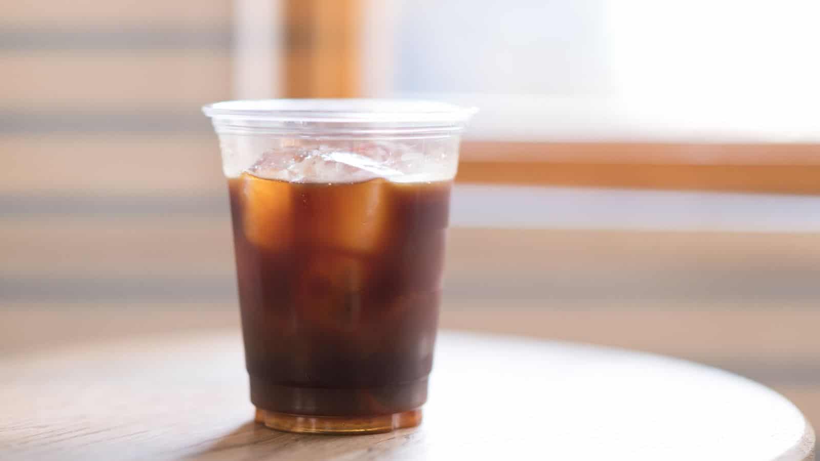 A plastic cup filled with iced coffee sits on a wooden table. The cup contains ice cubes and is placed near a window, allowing natural light to illuminate the beverage. The background is softly blurred.