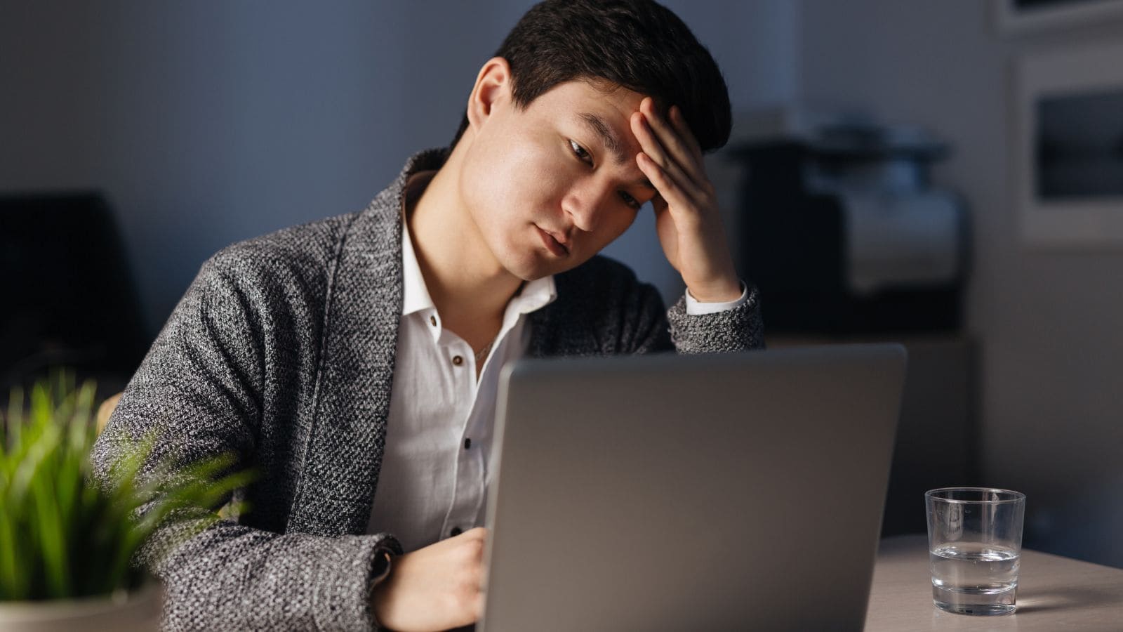 A person sits at a desk, looking at a laptop with a hand on their forehead. A glass of water and a small plant are on the desk nearby. The background is softly lit.