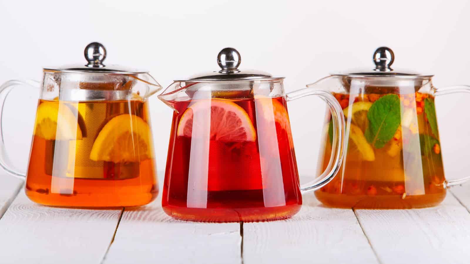 Three glass teapots filled with different types of tea are on a white wooden surface. Each teapot contains a metal lid and has slices of lemon and other ingredients visible inside, giving each a unique color and appearance.