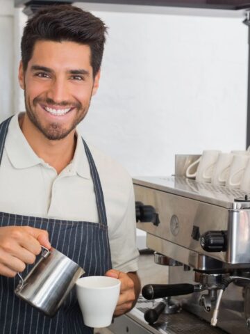 A person wearing an apron is smiling while preparing a coffee drink using an espresso machine. Stacked cups are visible on the machine, and a coffee grinder is nearby. The setting appears to be a café or coffee shop.