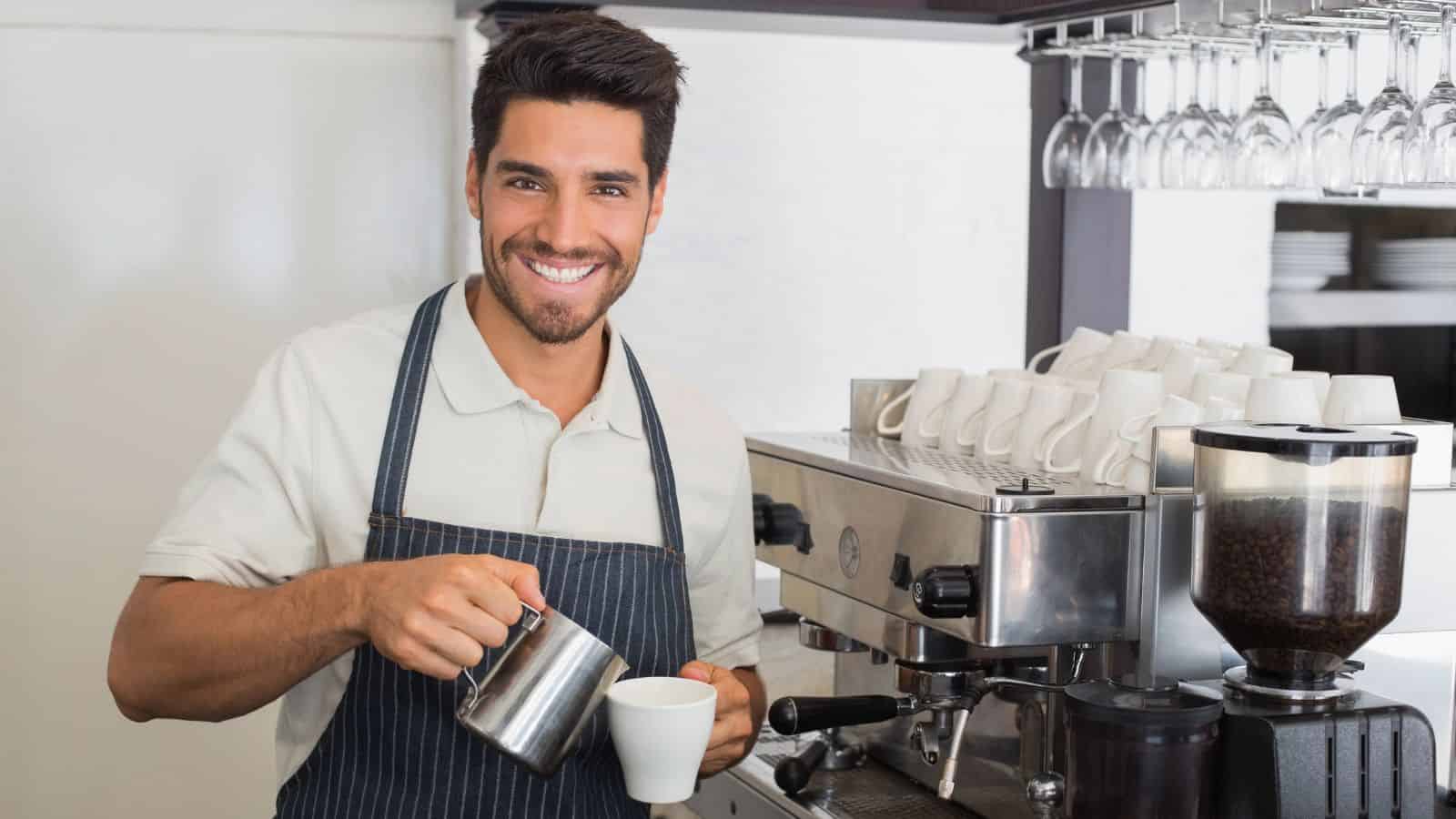 A person wearing an apron is smiling while preparing a coffee drink using an espresso machine. Stacked cups are visible on the machine, and a coffee grinder is nearby. The setting appears to be a café or coffee shop.