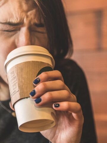 A person with shoulder-length brown hair is holding a coffee cup with a cardboard sleeve, squinting their eyes tightly shut. They are wearing a dark sweater, and the background is a wooden wall.