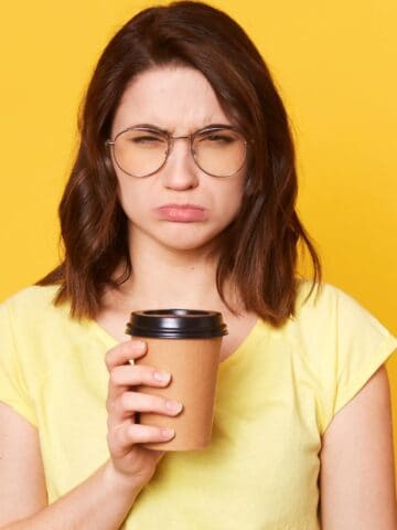 A person with shoulder-length brown hair and glasses frowns while holding a take-out coffee cup. They wear a yellow shirt and stand against a bright yellow background.