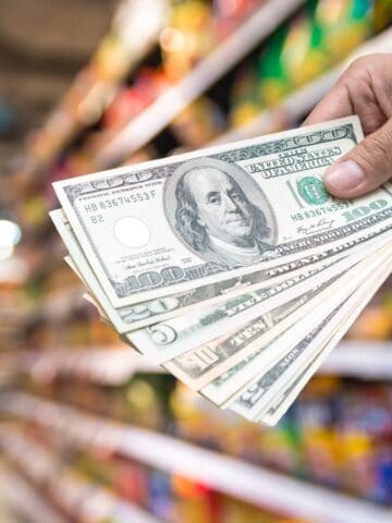 A hand holding several U.S. hundred-dollar bills in a grocery store aisle, with shelves stocked with various colorful products blurred in the background.