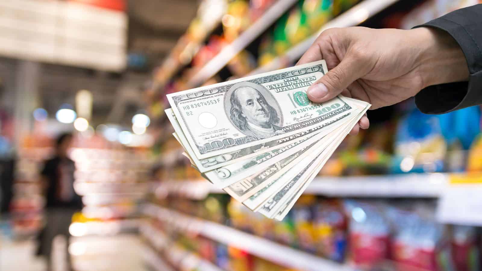 A hand holding several U.S. hundred-dollar bills in a grocery store aisle, with shelves stocked with various colorful products blurred in the background.