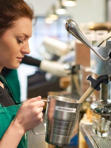 A barista in a black shirt and green apron is standing by a coffee machine, frothing milk in a metal pitcher. The background features shelves and another person in a similar uniform. The setting appears to be a busy café.