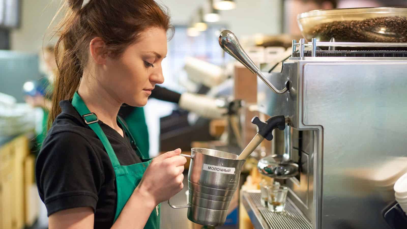 A barista in a black shirt and green apron is standing by a coffee machine, frothing milk in a metal pitcher. The background features shelves and another person in a similar uniform. The setting appears to be a busy caf&eacute;.