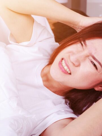 A person with long hair lies in bed under a white blanket, clutching their head with both hands and displaying a pained expression. They are wearing a white shirt, and sunlight is visible in the background.