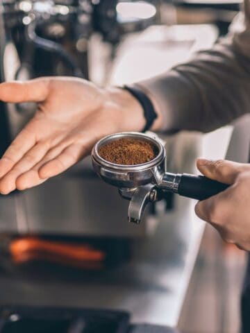 A person wearing a dark apron holds a portafilter filled with ground coffee in one hand and gestures with the other. Nearby, a coffee machine is partially visible, suggesting a coffee preparation setting.