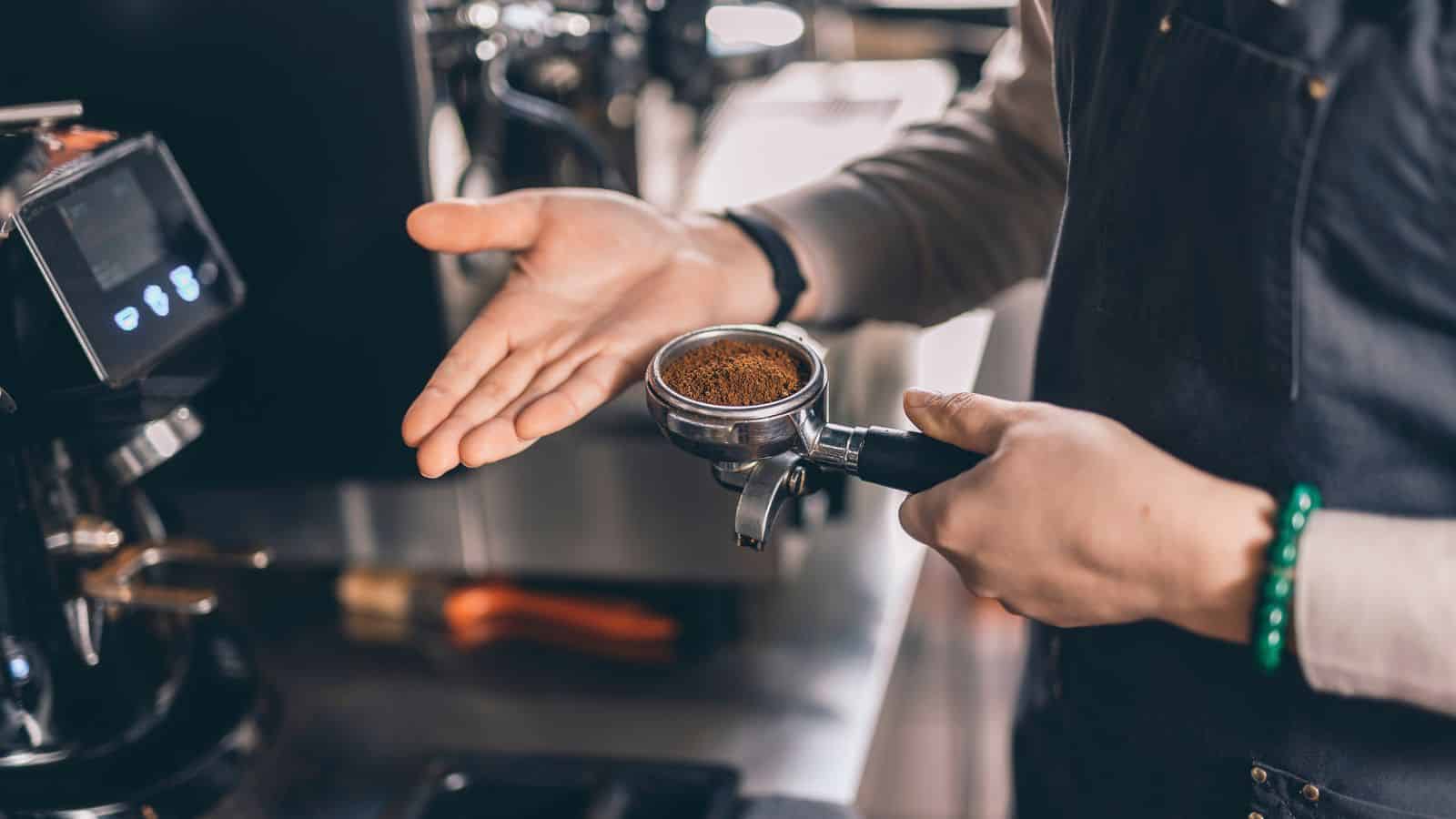 A person wearing a dark apron holds a portafilter filled with ground coffee in one hand and gestures with the other. Nearby, a coffee machine is partially visible, suggesting a coffee preparation setting.