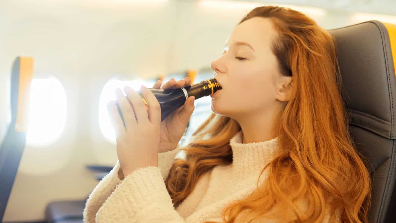 A person with long red hair is sitting in an airplane seat, drinking from a small bottle. They are wearing a white sweater and appear relaxed. The interior of the airplane is visible in the background.