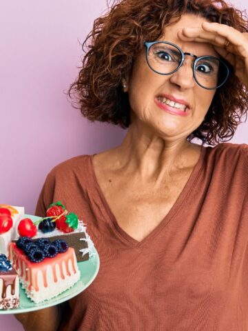A person with short curly hair and glasses holds a plate of assorted cakes, looking at the camera with a hand on their forehead. The background is a solid light pink color. They wear a brown V-neck shirt and a red bracelet.