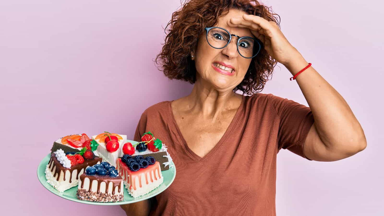 A person with short curly hair and glasses holds a plate of assorted cakes, looking at the camera with a hand on their forehead. The background is a solid light pink color. They wear a brown V-neck shirt and a red bracelet.