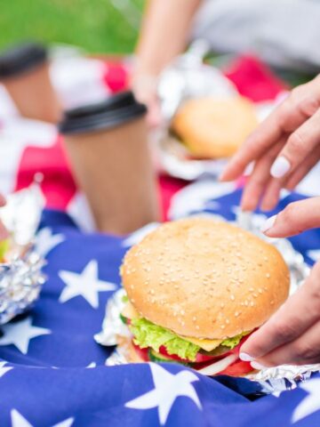 Hands reaching for burgers wrapped in foil, placed on an American flag laid on grass. Disposable coffee cups are also visible.