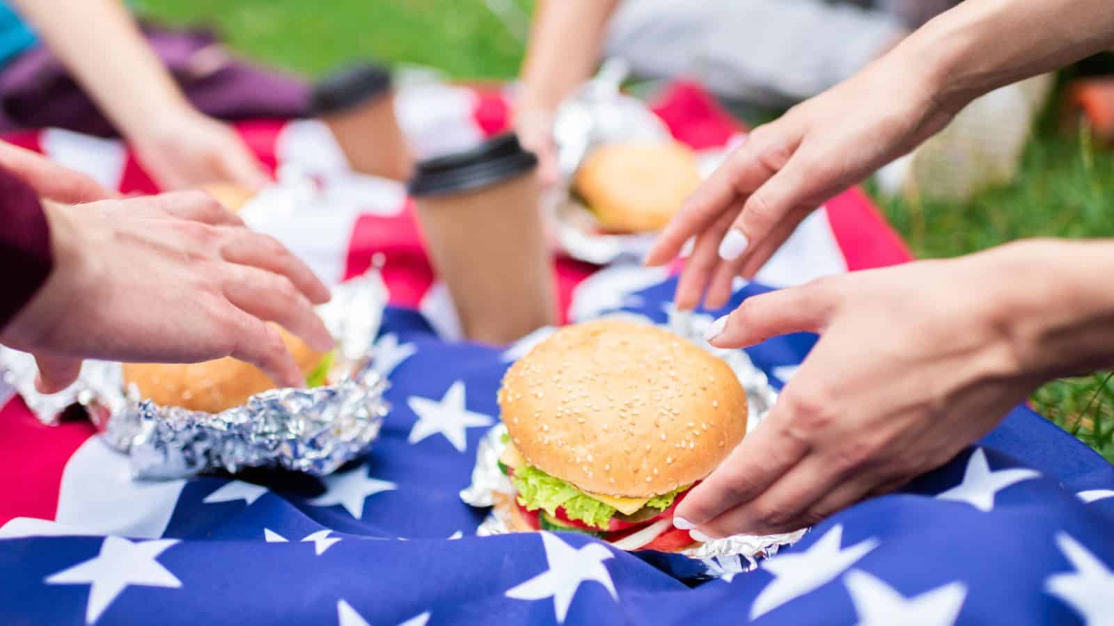 Hands reaching for burgers wrapped in foil, placed on an American flag laid on grass. Disposable coffee cups are also visible.