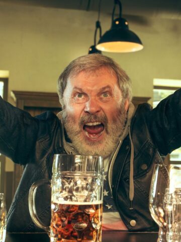 A bearded man in a denim jacket is at a bar with his arms raised and mouth open. A large beer mug and several glasses are on the counter in front of him. Hanging lamps provide warm lighting.