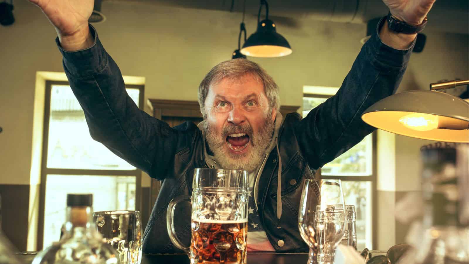 A bearded man in a denim jacket sits at a bar, raising his arms enthusiastically. In front of him are a few glasses, including a large mug of beer. The background shows hanging lights and windows.