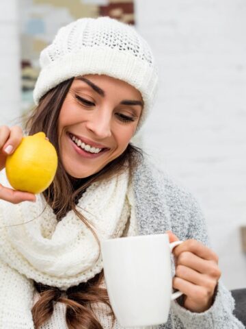 A woman wearing a knit hat and scarf sits in a cozy room, smiling as she holds a yellow lemon above a white mug. The background features a gray couch, shelves, and a desk with a laptop.