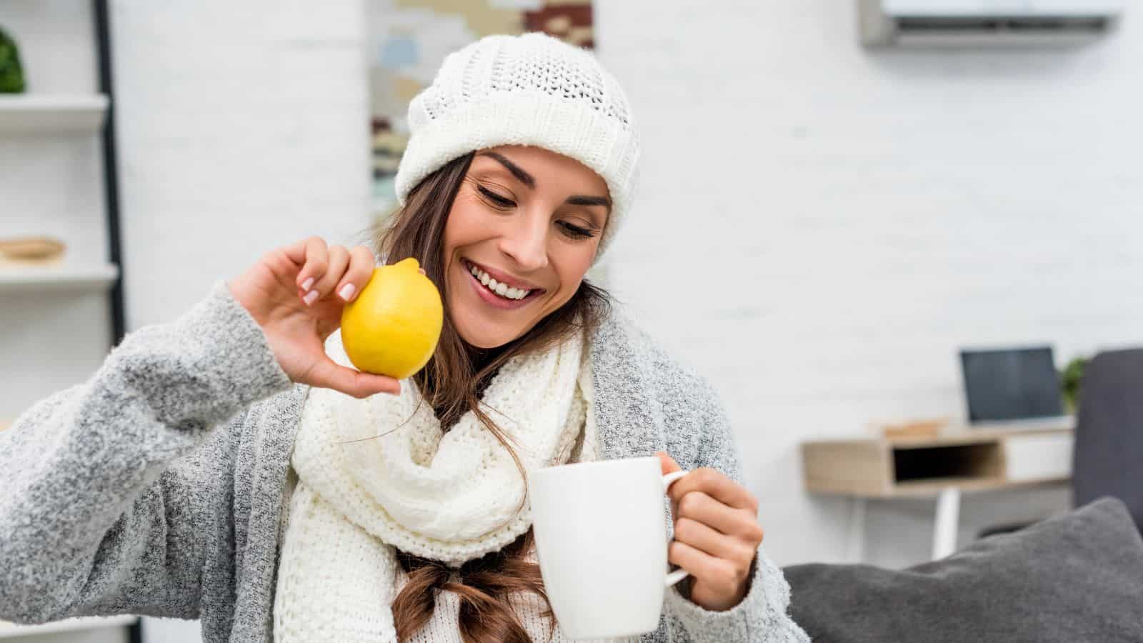 A woman wearing a knit hat and scarf sits in a cozy room, smiling as she holds a yellow lemon above a white mug. The background features a gray couch, shelves, and a desk with a laptop.