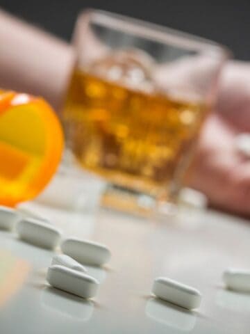 A close-up of a hand resting near a spilled prescription bottle with white pills scattered on a surface. A glass of amber liquid is nearby, suggesting alcohol. The scene implies a cautionary situation regarding substance use or overdose.