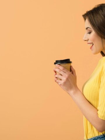 A woman wearing a yellow shirt is holding a takeout coffee cup. She is smiling and looking at the cup. The background is a solid peach color.