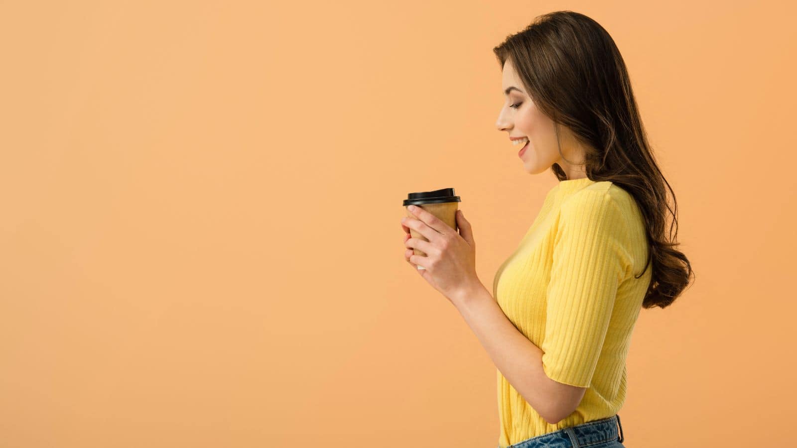 A woman wearing a yellow shirt is holding a takeout coffee cup. She is smiling and looking at the cup. The background is a solid peach color.