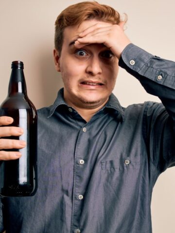 A man with a surprised expression holds a dark bottle in one hand and touches his forehead with the other. He is wearing a gray button-up shirt and stands against a plain beige background.