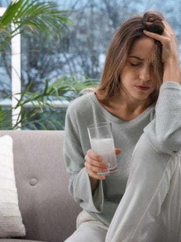 A woman sits on a gray sofa holding her head with one hand and a glass of milk in the other. She appears distressed. A pillow is beside her, and a large window with a view of a snowy landscape is in the background.