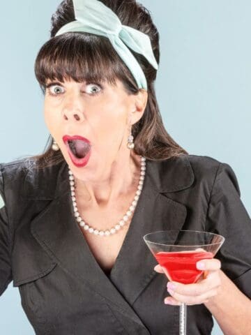 A woman with a surprised expression holds a retro telephone to her ear and a martini glass filled with a red drink in her other hand. She is dressed in vintage attire with a black dress, pearl necklace, and a light blue headband.