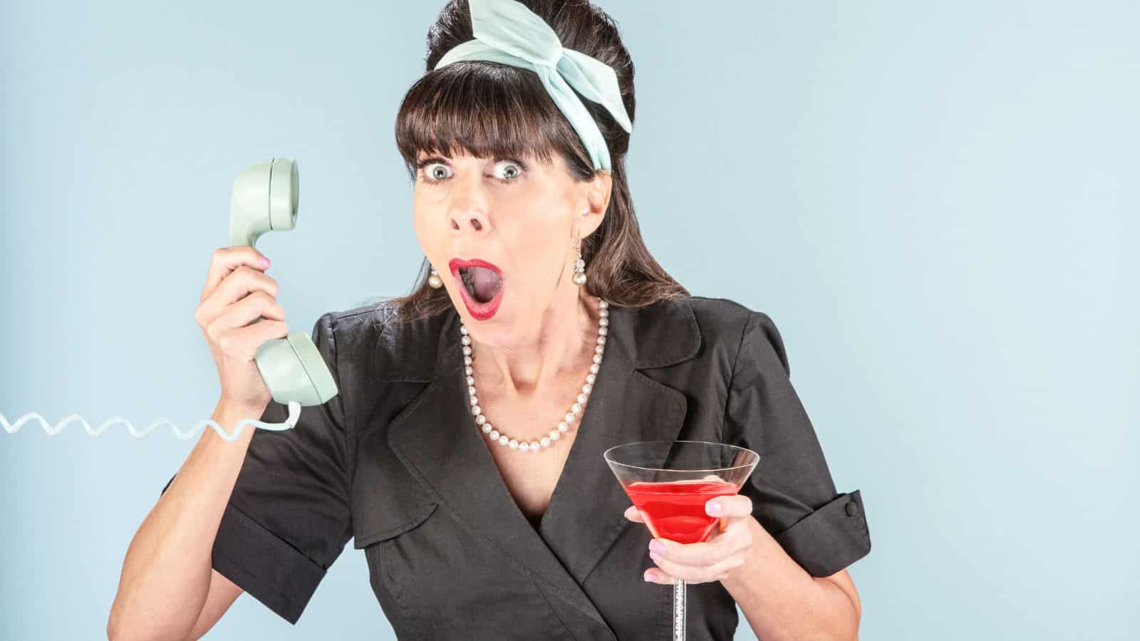 A woman with a surprised expression holds a retro telephone to her ear and a martini glass filled with a red drink in her other hand. She is dressed in vintage attire with a black dress, pearl necklace, and a light blue headband.