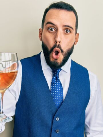 A man with a beard and mustache, wearing a blue vest and polka dot tie, holds a large wine glass. He has a surprised expression and is standing against a plain light-colored background.