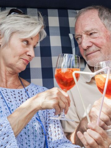 An older couple sits together in a checkered outdoor seating area, holding cocktails with orange slices and white straws. They are clinking glasses and smiling at each other, wearing casual clothing in a relaxed setting.