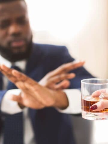 A man in a suit sitting at a table gestures with his hands to refuse a glass of dark liquid being offered to him by another person. The background is out of focus, suggesting an indoor setting with natural light.