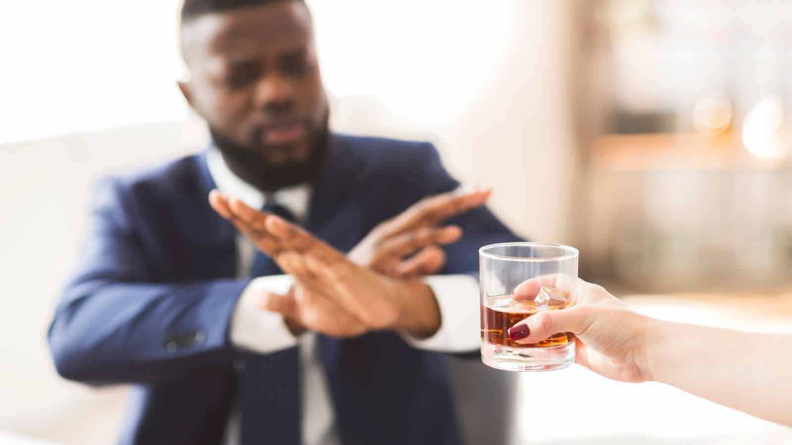 A man in a suit sitting at a table gestures with his hands to refuse a glass of dark liquid being offered to him by another person. The background is out of focus, suggesting an indoor setting with natural light.