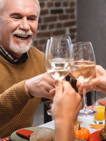 A group of three people sit at a table, toasting with glasses of white wine. An older man with a beard smiles while holding his glass. There is a roasted turkey on the table, along with decorative pumpkins and a festive setting.