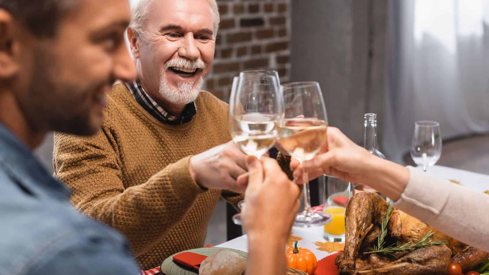 A group of three people sit at a table, toasting with glasses of white wine. An older man with a beard smiles while holding his glass. There is a roasted turkey on the table, along with decorative pumpkins and a festive setting.