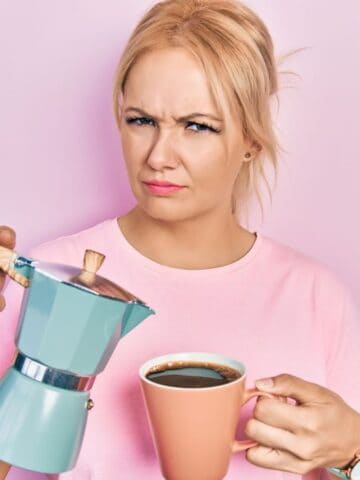 A person with blonde hair wearing a pink shirt is holding a blue coffee pot and a pink coffee cup. They have a puzzled expression and are standing against a light pink background.