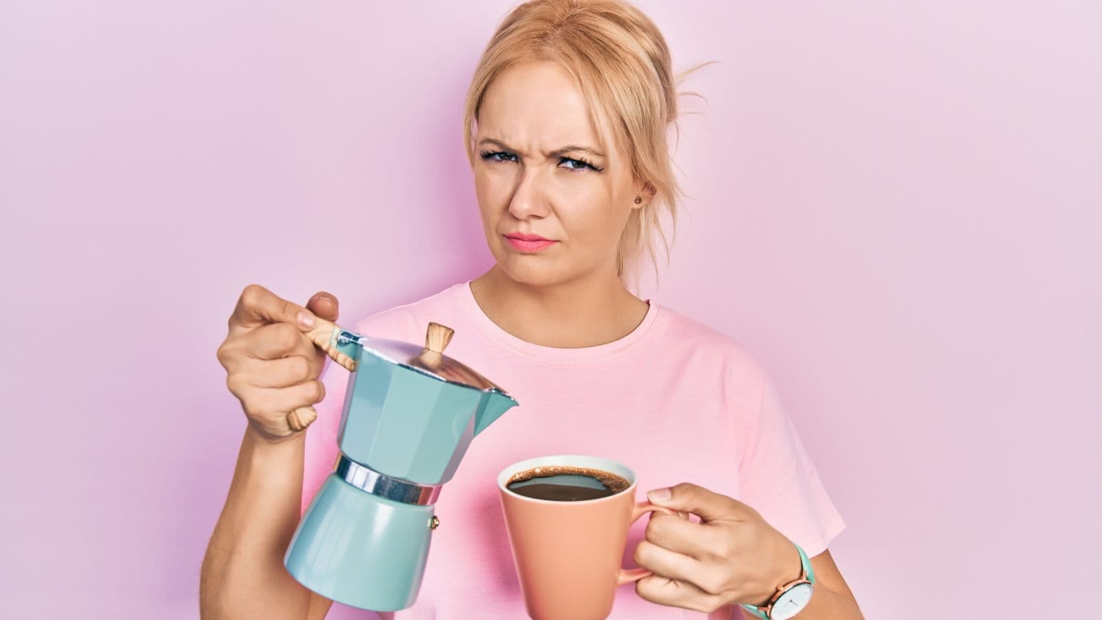 A person with blonde hair wearing a pink shirt is holding a blue coffee pot and a pink coffee cup. They have a puzzled expression and are standing against a light pink background.