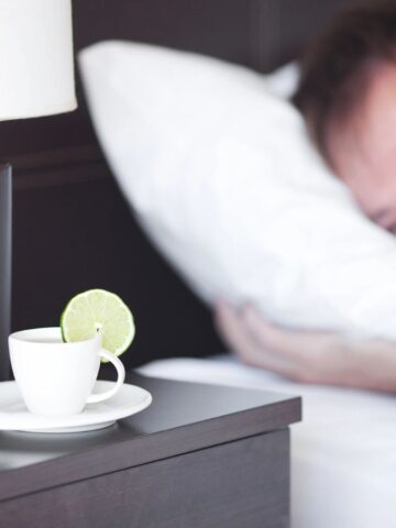 A man lies in bed with a smile, resting his head on a pillow. On the bedside table, there is a white cup and saucer with a lime slice on the rim. Beside it is a lamp with a white shade and a dark base.