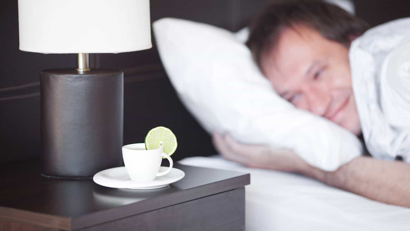 A man lies in bed with a smile, resting his head on a pillow. On the bedside table, there is a white cup and saucer with a lime slice on the rim. Beside it is a lamp with a white shade and a dark base.