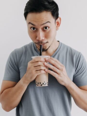 A person with short hair, wearing a light gray t-shirt, drinks from a plastic cup with a black straw. The cup contains a brown beverage with black tapioca pearls at the bottom. The background is plain white.