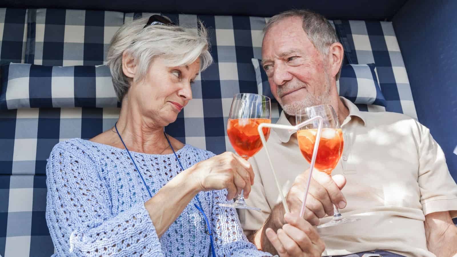 An older couple sits together in a checkered outdoor seating area, holding cocktails with orange slices and white straws. They are clinking glasses and smiling at each other, wearing casual clothing in a relaxed setting.