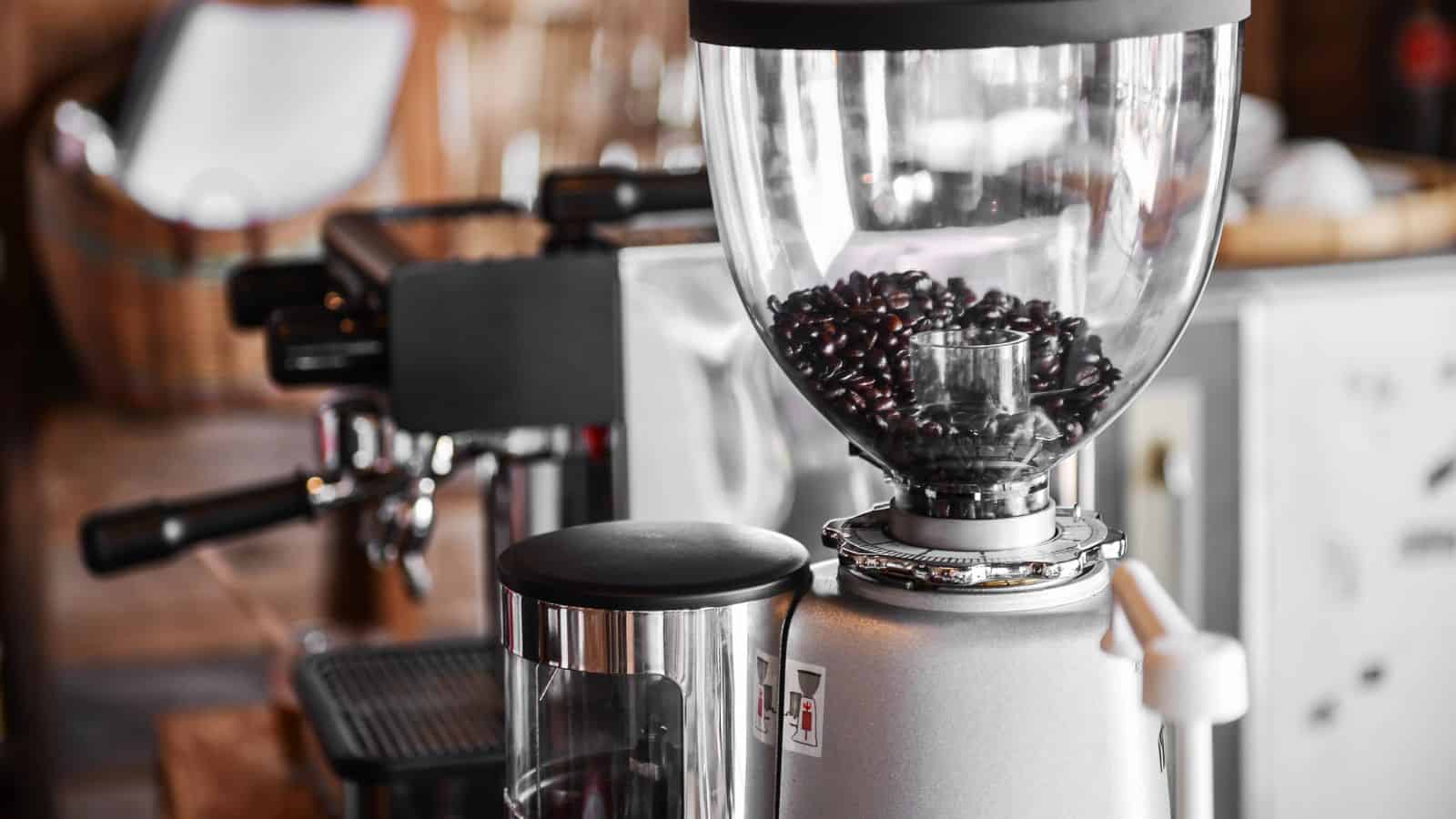 A close-up of a coffee grinder filled with dark coffee beans. The machine is metallic with a transparent hopper. In the background, a blurred coffee machine and other kitchen items are visible.