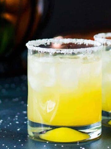 Two glasses of yellow cocktail with ice and salted rims are placed on a dark surface. A wedge of lime is inside each glass. There are scattered salt crystals around the glasses. A blurred background shows brown and green objects.